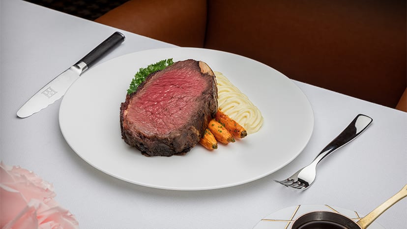 A slice of prime rib sits on baby carrots on a white plate.