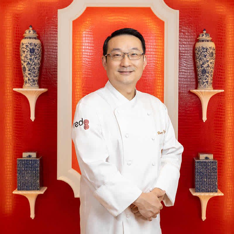 Chef Kevin HanQing Yuan stands in from of a red wall with vases at Encore Boston Harbor